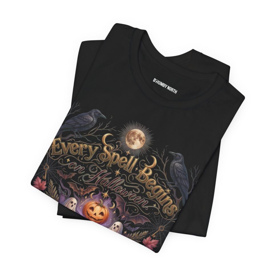 Woman wearing a black Bella+Canvas Halloween graphic t-shirt featuring a vintage illustration of an enchanted spell book, pumpkin, ghosts, candles, ravens, and the phrase "Every Spell Begins on Halloween."