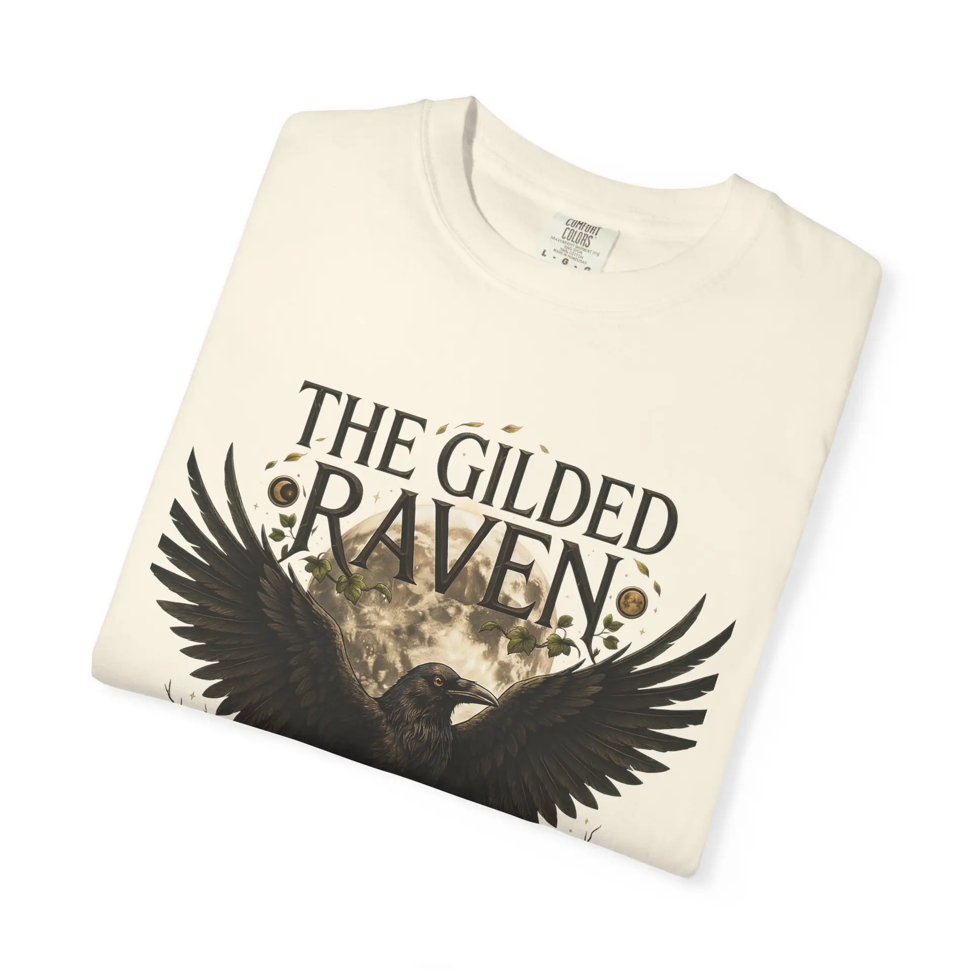 Person wearing a The Gilded Raven graphic t-shirt featuring a minimalist raven positioned before a glowing full moon with bold vintage-inspired typography reading "The Gilded Raven," photographed in a professional studio.