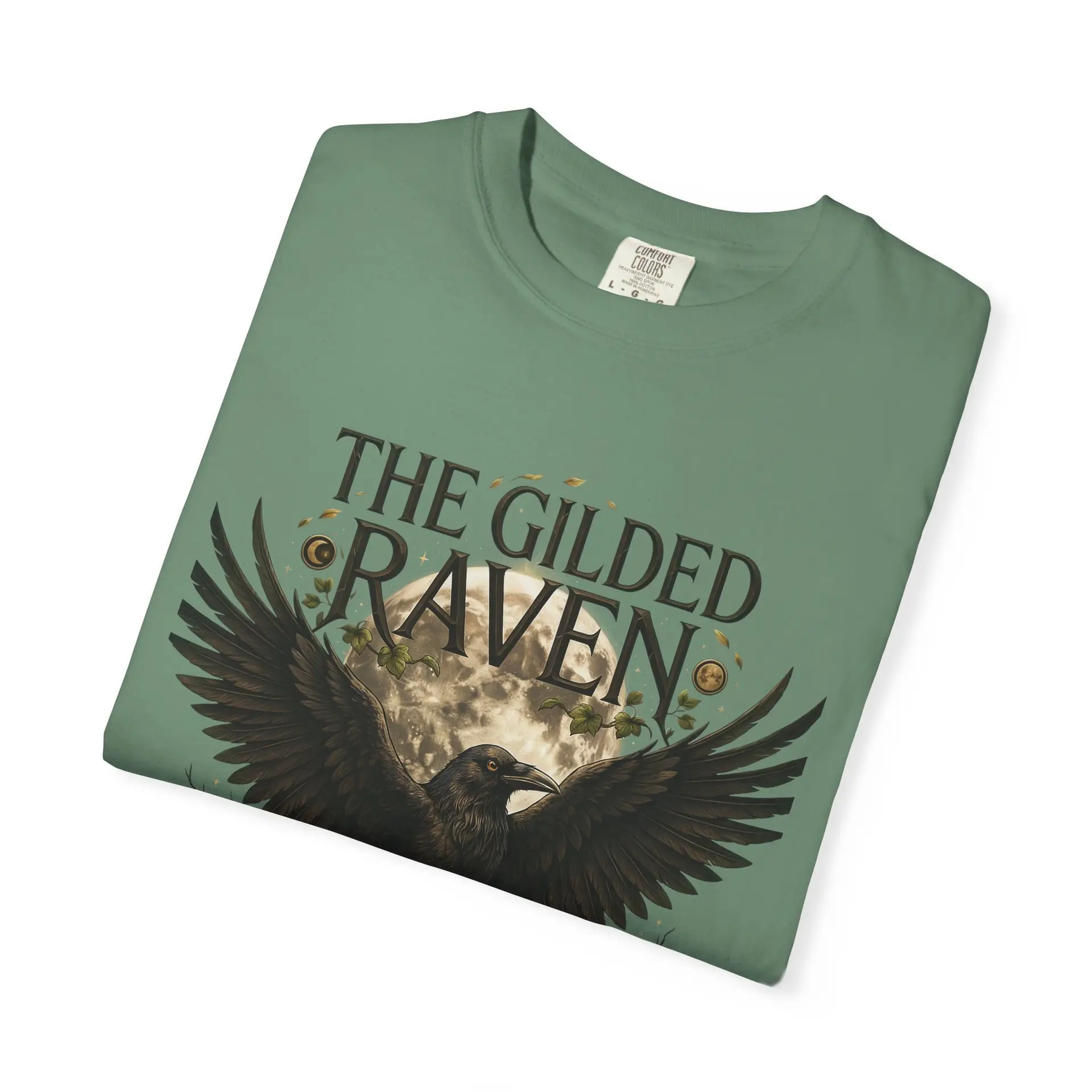 Person wearing a The Gilded Raven graphic t-shirt featuring a minimalist raven positioned before a glowing full moon with bold vintage-inspired typography reading "The Gilded Raven," photographed in a professional studio.