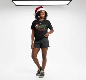 Smiling woman wearing a black Merry Christmas plaid graphic unisex t-shirt featuring festive lettering, Santa hat, reindeer antlers, and holiday decorations in a white studio setting.