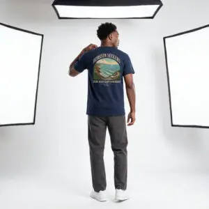 Someone wearing a blue Horizon Seekers graphic t-shirt featuring a vintage coastal road illustration with ocean cliffs, a retro car, bold typography reading "Every Road Leads Somewhere," and a nostalgic travel-inspired design in a professional studio.