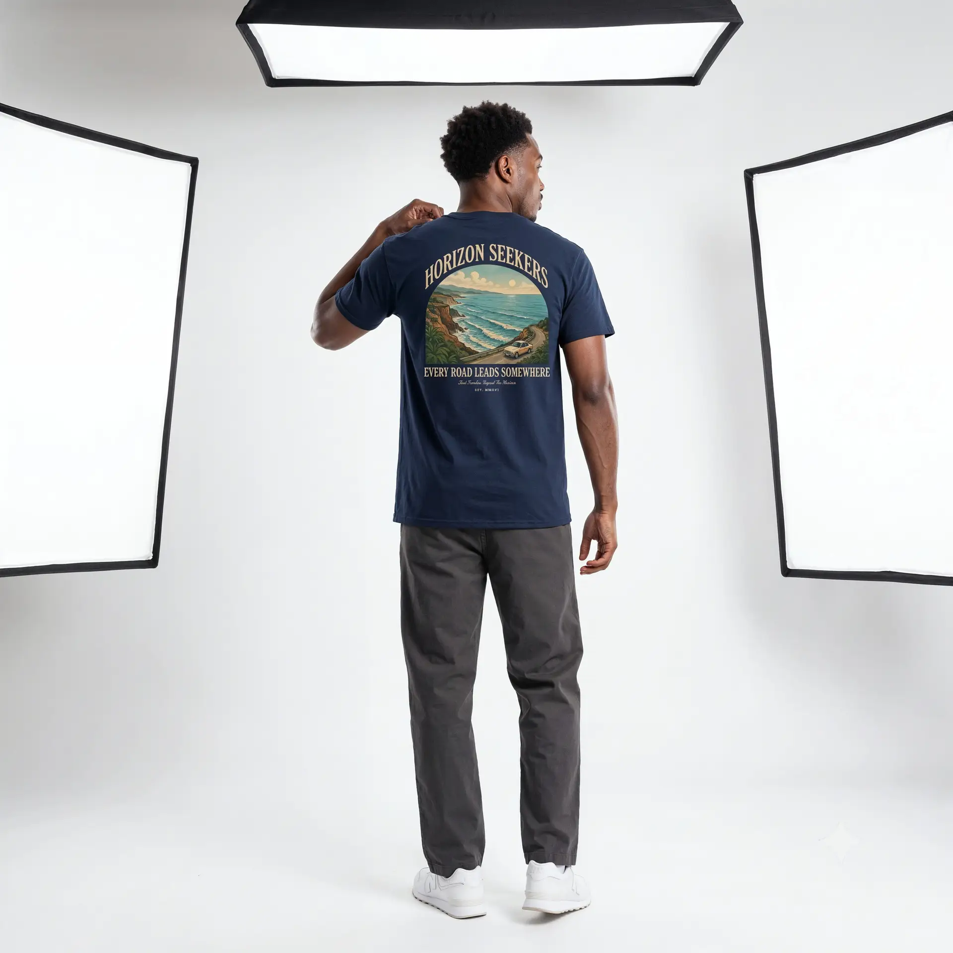 Someone wearing a blue Horizon Seekers graphic t-shirt featuring a vintage coastal road illustration with ocean cliffs, a retro car, bold typography reading "Every Road Leads Somewhere," and a nostalgic travel-inspired design in a professional studio.