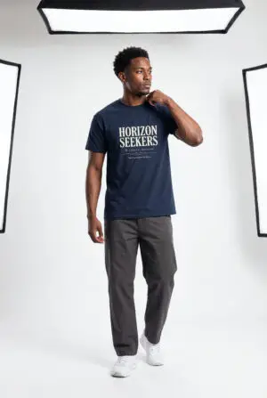 Someone wearing a blue Horizon Seekers graphic t-shirt featuring a vintage coastal road illustration with ocean cliffs, a retro car, bold typography reading "Every Road Leads Somewhere," and a nostalgic travel-inspired design in a professional studio.