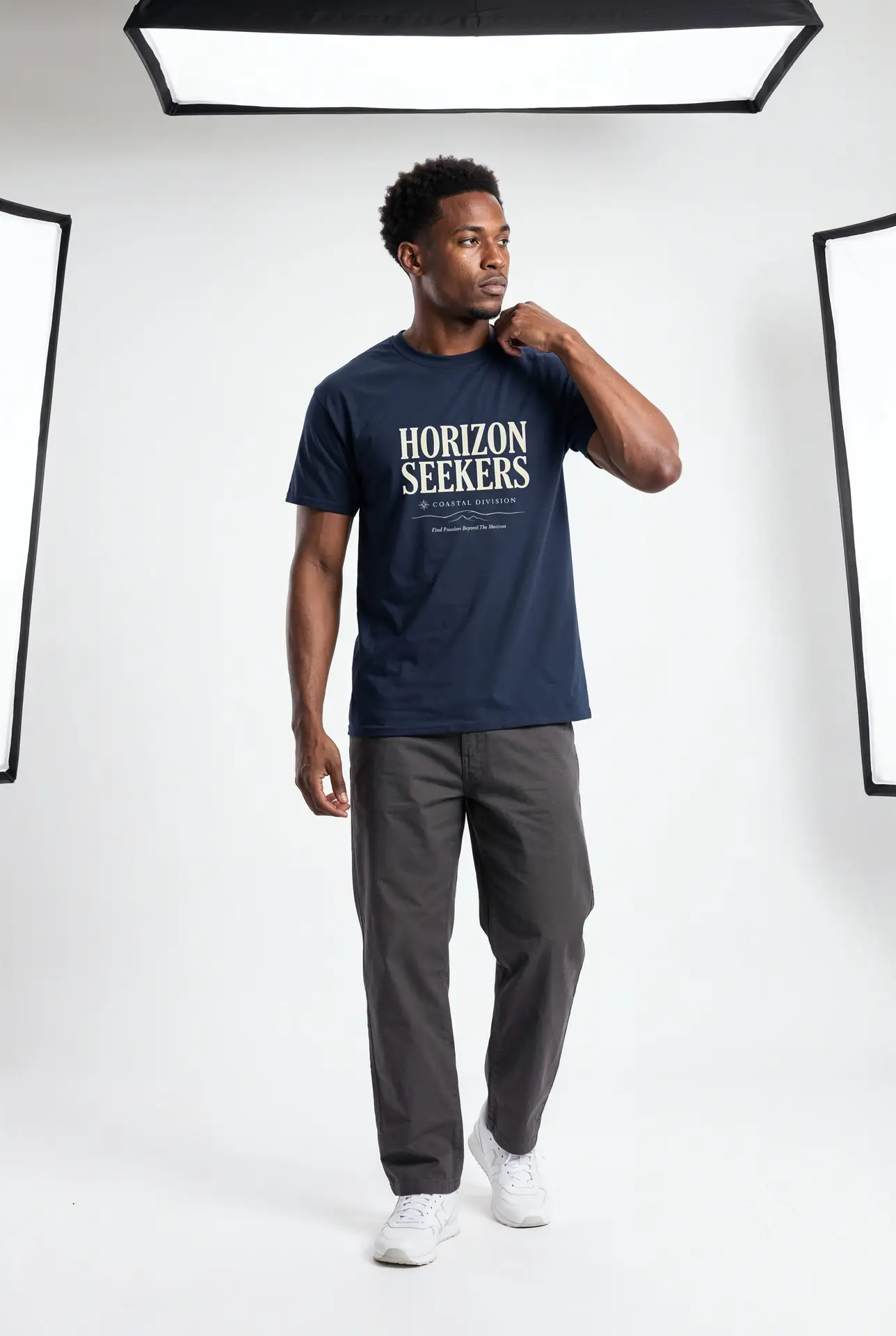 Someone wearing a blue Horizon Seekers graphic t-shirt featuring a vintage coastal road illustration with ocean cliffs, a retro car, bold typography reading "Every Road Leads Somewhere," and a nostalgic travel-inspired design in a professional studio.