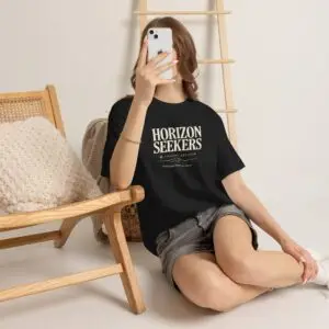 Someone wearing a blue Horizon Seekers graphic t-shirt featuring a vintage coastal road illustration with ocean cliffs, a retro car, bold typography reading "Every Road Leads Somewhere," and a nostalgic travel-inspired design in a professional studio.