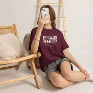 Someone wearing a blue Horizon Seekers graphic t-shirt featuring a vintage coastal road illustration with ocean cliffs, a retro car, bold typography reading "Every Road Leads Somewhere," and a nostalgic travel-inspired design in a professional studio.
