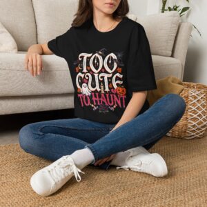 Woman wearing a black Too Cute To Haunt Halloween graphic t-shirt featuring vintage typography, a cute ghost, pumpkin, autumn leaves, and spooky accents in a professional studio.
