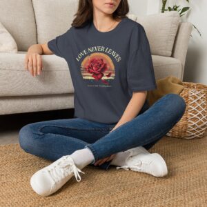 Person wearing a Love Never Leaves graphic t-shirt featuring a vintage-inspired bouquet of deep red roses, warm sunset tones, scattered rose petals, and elegant typography reading "Love Never Leaves" and "Only The Moment Does," photographed in a professional studio.
