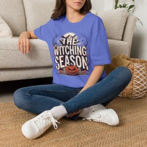 Woman wearing a blue The Witching Season Halloween graphic t-shirt featuring vintage spooky typography, a smiling jack-o'-lantern, moon, and autumn tree silhouettes in a professional studio.