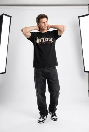 Woman wearing a black Mistletoe Social Club Christmas graphic t-shirt featuring vintage holiday typography with festive mistletoe accents