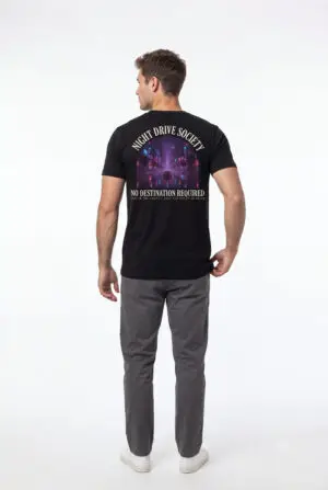 Person wearing a Night Drive Society graphic t-shirt featuring a retro-futuristic neon city skyline, rain-soaked streets, towering palm trees, and a classic car driving into the night with bold typography reading "No Destination Required" and "Lost In The Lights • Built For The After Hours," photographed in a professional studio.