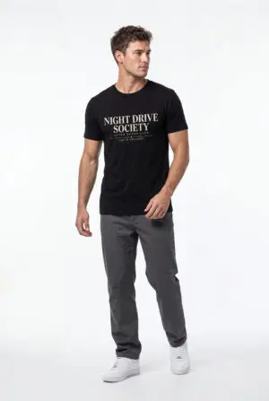 Person wearing a Night Drive Society graphic t-shirt featuring a retro-futuristic neon city skyline, rain-soaked streets, towering palm trees, and a classic car driving into the night with bold typography reading "No Destination Required" and "Lost In The Lights • Built For The After Hours," photographed in a professional studio.