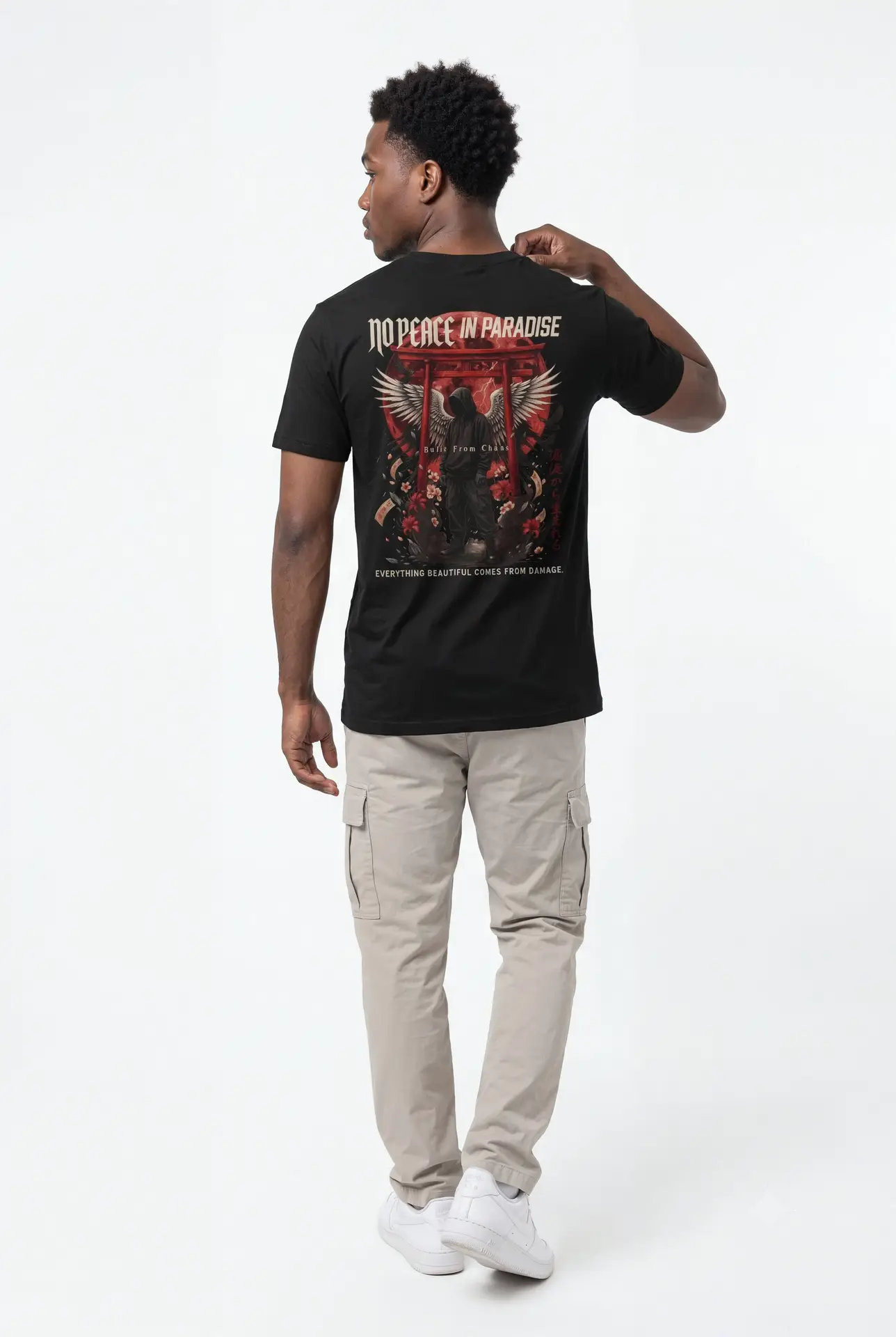 Person wearing a black No Peace In Paradise graphic t-shirt featuring a hooded figure with angel wings beneath a traditional Japanese torii gate, set against a crimson moon with ravens, floral elements, and gothic typography reading "Built From Chaos" and "Everything Beautiful Comes From Damage," photographed in a professional studio.