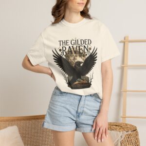 Person wearing a The Gilded Raven graphic t-shirt featuring a minimalist raven positioned before a glowing full moon with bold vintage-inspired typography reading "The Gilded Raven," photographed in a professional studio.