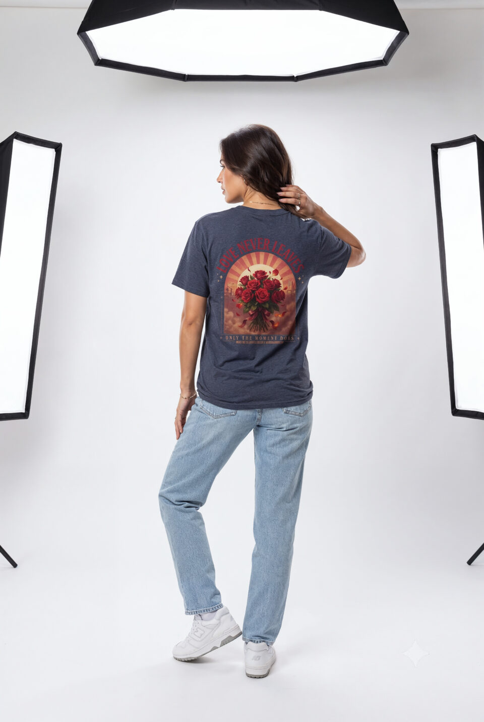 Person wearing a Love Never Leaves graphic t-shirt featuring a vintage-inspired bouquet of deep red roses, warm sunset tones, scattered rose petals, and elegant typography reading "Love Never Leaves" and "Only The Moment Does," photographed in a professional studio.