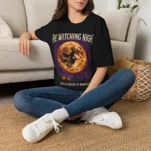 Front view of a black Gildan Softstyle 64000 unisex t-shirt featuring "The Witching Night" vintage Halloween graphic with a full moon, flying witch, and gothic typography, modeled in a studio setting.