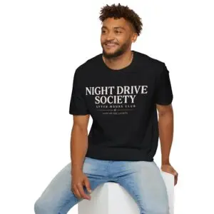 Person wearing a black Night Drive Society graphic t-shirt featuring a retro-futuristic neon city skyline, rain-soaked streets, towering palm trees, and a classic car driving into the night with bold typography reading "No Destination Required" and "Lost In The Lights • Built For The After Hours," photographed in a professional studio.